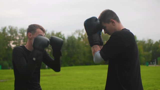 white men playful sparring in park, lighthearted mitt exchanges and smiling jabs between friends casual fitness session with overcast sky, easy pace, banter, coaching tips, and warm energy captured