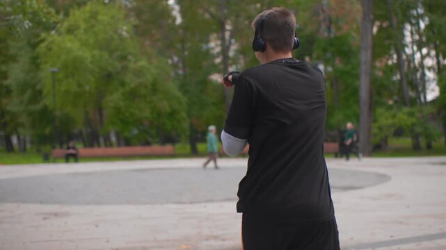 caucasian man shadowboxing in park headphones on, fingerless gloves and visible tattoo, focused punches and quick footwork on circular plaza, trees and benches in background, intense urban fitness