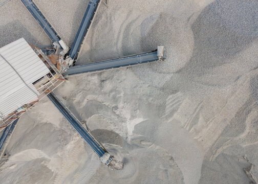 Aerial View of Rock Crusher Machinery and a Conveyor Belt System Near Large Aggregate Stockpiles at an Industrial Quarry Site. Rock Crushing and Aggregate Production Viewed from Above.