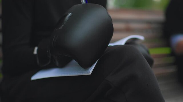 Black boxing gloves on lap caucasian man quietly sketching tactics on paper while seated on wooden park bench, closeup of leather mitt and pen, muted daylight, textured clothing, contemplative mood