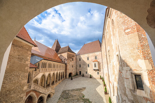 Corvin Castle courtyard framing the elaborate gothic architecture, historic walls, and red-tiled roofs against a cloudy sky, showing centuries of history in Transylvania, Romania