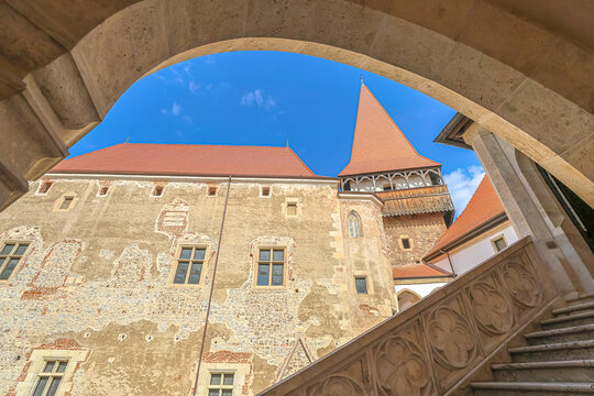 Corvin Castle courtyard view framed by a gothic arch, revealing terracotta roofs, aged stone walls and a pointed tower with decorative staircase under a clear blue sky in Transylvania, Romania