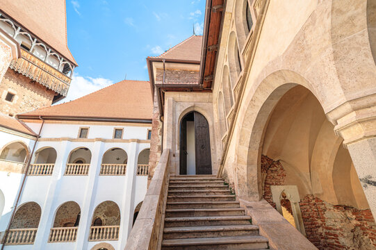 Corvin Castle courtyard in Hunedoara showcases Gothic-Renaissance stone steps, arched doorway and balustrades beneath a clear blue sky, a historic Transylvanian landmark in Romania