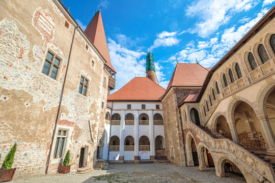 Corvin Castle in Hunedoara, Romania, showing its historical courtyard with intricate stone carvings, pointed arches, and varying rooflines under a bright summer sky in Transylvania