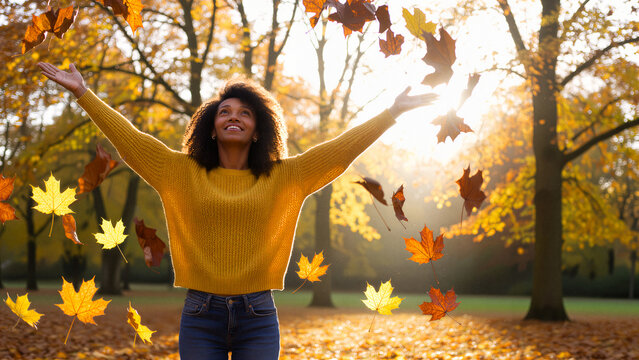 Casual joyful black woman having fun throwing leaves and raising her arms in autumn at city park.