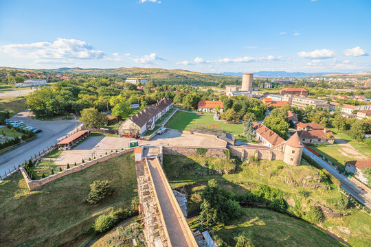 Hunedoara cityscape showcasing residential buildings, green areas, roads, and a bridge leading towards the historic Corvin Castle, viewed from above under a clear blue sky in Transylvania, Romania