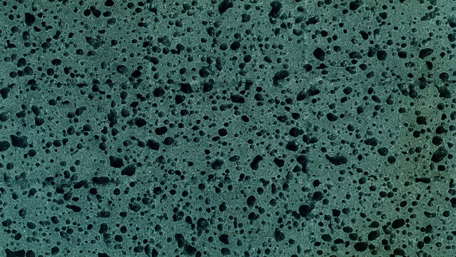A close-up texture of a porous material in a turquoise or deep greenish-blue shade, featuring a pitted surface reminiscent of foam rubber or pumice.