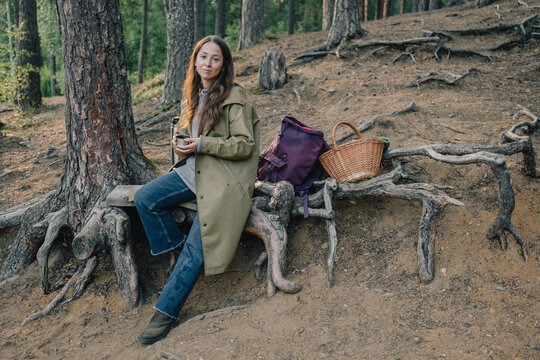 Woman resting with a hot drink beside a pine tree in the forest, with backpack and wicker basket, enjoying a peaceful break during hiking and seasonal foraging.