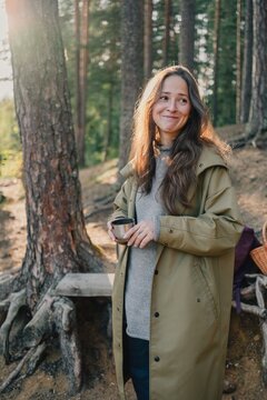 Smiling woman holding a metal cup in a sunlit pine forest, enjoying a peaceful outdoor break, warm drink, nature retreat and restorative escape from city life