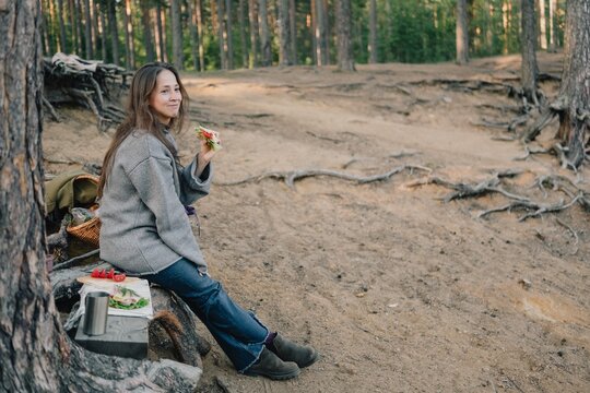 Woman sitting on exposed tree roots and eating a fresh sandwich during a forest picnic, enjoying a relaxed hiking break, outdoor meal and peaceful nature escape.
