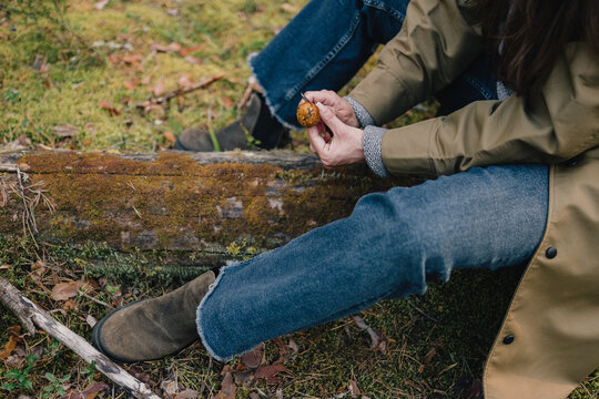 Woman examining a freshly picked wild mushroom while resting on a mossy log in the forest, illustrating mushroom foraging, nature exploration and outdoor lifestyle.