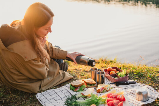 Woman pouring a hot drink from a thermos during a lakeside picnic at golden hour, with fresh sandwiches, fruit and vegetables, enjoying relaxed outdoor leisure.