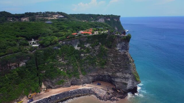 Pura Luhur Uluwatu temple complex sits on a high cliff above active coastal construction works and calm tropical sea.
