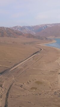 Drone footage of the Orto-Tokoy Reservoir with a winding shoreline road, frozen water edge and barren steppe landscape, framed by snowcapped mountains, Kyrgyzstan