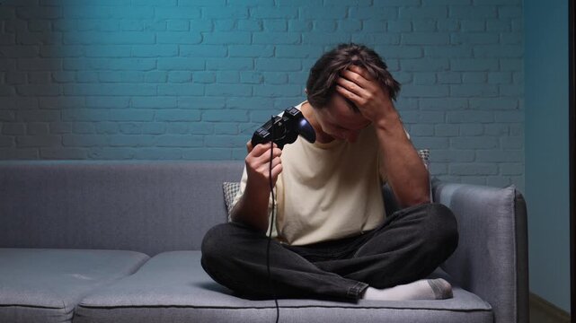 Young man experiencing frustration and disappointment after losing a video game, sitting cross legged on a gray sofa in a dimly lit room with a blue brick wall