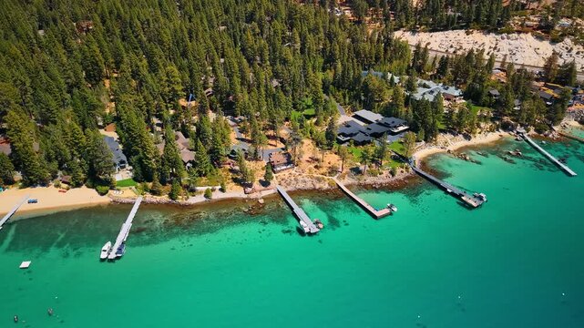 Waterfront homes and piers among conifers at Lake Tahoe. Docks extend from a narrow beach into turquoise shallows below houses and a dense canopy of trees.
