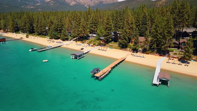 Piers extending from a sandy beach at Lake Tahoe. Docks cast shadows into green water below a conifer shoreline, with rows of loungers along the sand.