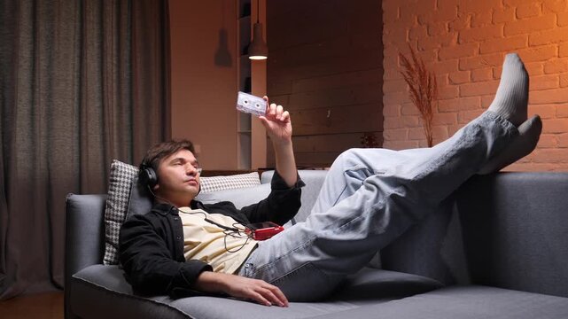 Young man wearing headphones lying on a sofa, holding an old cassette tape, enjoying vintage music, evoking feelings of nostalgia and comfort in a cozy home setting