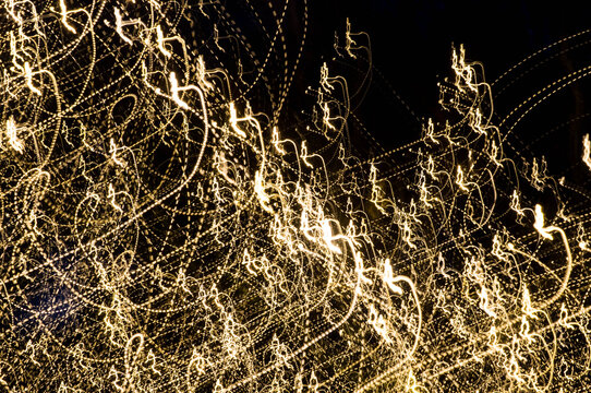 A turbulent mass of swirling and dotted golden light paths. Distant darker tree silhouettes add depth to the abstract Christmas light scene captured with long exposure.