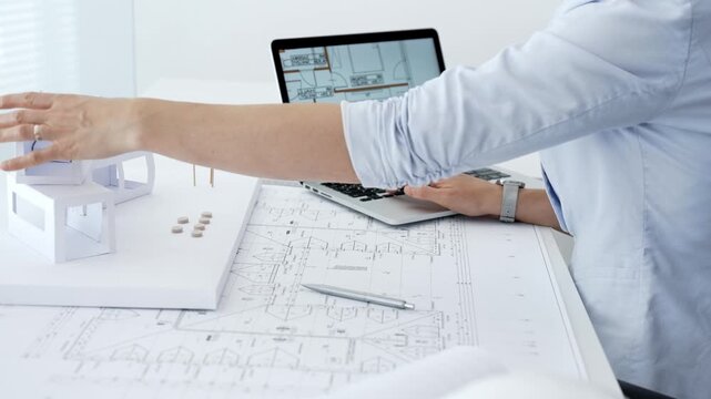 Architect actively building a paper architectural model on a desk strewn with blueprints and a laptop showing digital floor plans, capturing hands on design process