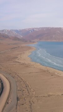 Drone footage of the Orto-Tokoy Reservoir with turquoise water, sandy shoreline, and winding road, framed by snowcapped mountains in Issyk-Kul Region, Kyrgyzstan