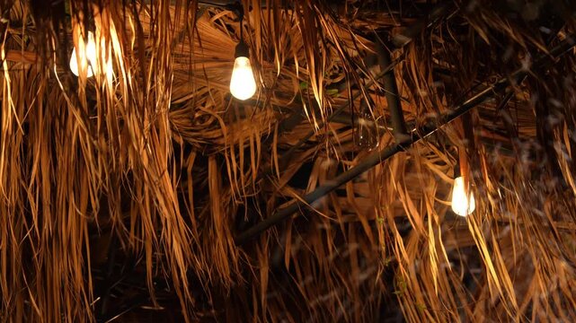 Warm bulb string lights hang beneath a straw thatched roof, swaying and flickering as strong wind and an approaching storm shake the dried palm leaves at night.