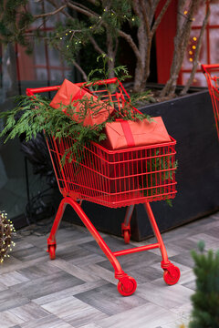 Red shopping cart filled with wrapped Christmas gifts and fresh evergreen branches, festive holiday decoration outside a store, colorful seasonal display creating a cheerful Christmas shopping atmosph