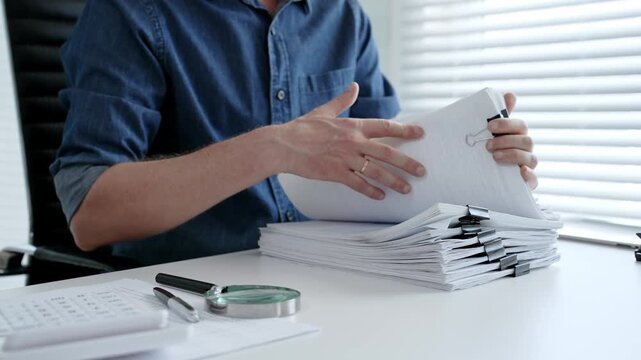 Person's hands organizing a large stack of office documents, checking paperwork for accuracy, analyzing data, and managing files on a desk