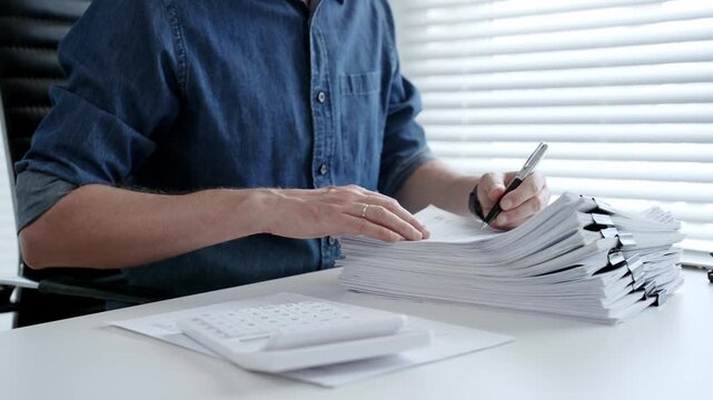 Individual working diligently on financial documents, calculating expenses and reviewing reports at an office desk, managing a large stack of paperwork and invoices