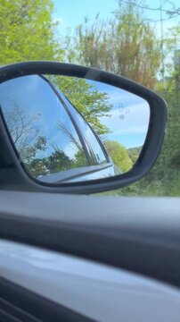 Side view mirror of a car reflecting the scenic country road and lush green forest during a summer road trip, showcasing the blue sky and trees passing by from the passenger's perspective