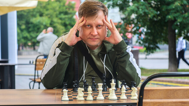 Adult man sitting outdoor chessboard in a park, deeply thinking and strategizing his next move in a classic intellectual board game.