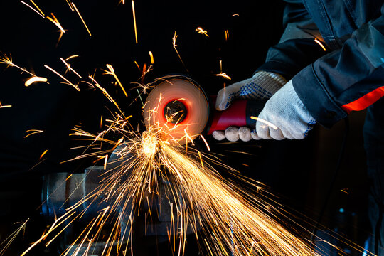 A worker in protective gloves uses an angle grinder to cut metal clamped in a heavy bench vice. Intense stream of bright orange sparks flying in a dark workshop, industrial metalworking manufacturing 