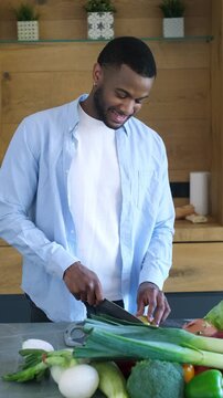 Smiling young african american man standing in a modern kitchen, using a sharp knife to slice fresh vegetables on a cutting board, preparing ingredients for a healthy home-cooked meal
