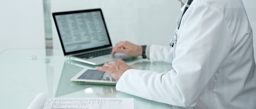 Healthcare professional using a laptop, digital tablet, and medical records, managing patient information and health data in a modern clinic or hospital setting