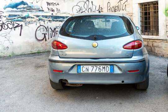 Barletta, Italy - Aug 1, 2026: Silver Alfa Romeo 147 hatchback parked in a small square in front of a wall covered with graffiti tags and a painted sea mural