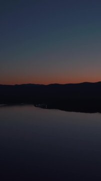 Aerial footage at dusk over Lake Turgoyak with smooth dark water, distant mountain silhouettes and a colorful sunset sky, tranquil nature landscape in Chelyabinsk Oblast, Russia