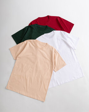 Flat lay of four colorful blank t shirts in beige white dark green and red arranged neatly on a light background for fashion and apparel catalog presentation
