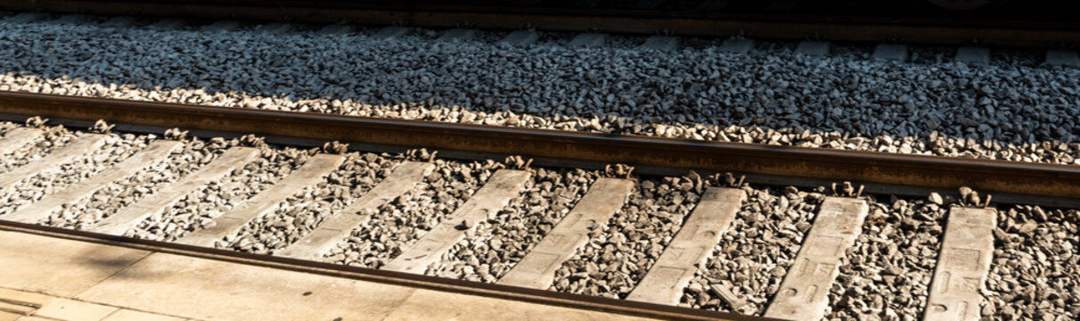 Wide panoramic strip of railway tracks with sleepers and crushed stone ballast lit by warm sunlight beside a platform edge, concept of train journey, rail infrastructure and travel background