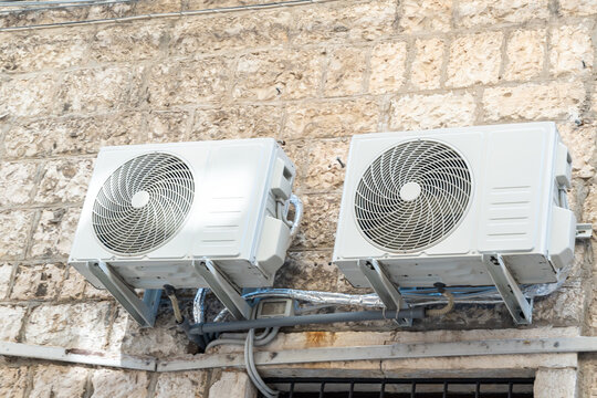 Two white air conditioning outdoor units mounted on metal brackets on an old limestone block wall with pipes and cables running below, concept of cooling in heatwave, energy consumption and home clim