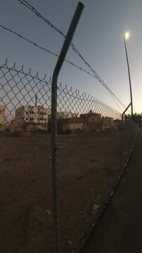 Tracking shot past a chain-link fence showing an empty lot and buildings at dusk.
