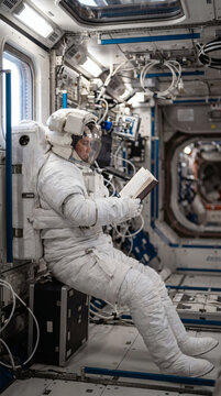 An astronaut in a full spacesuit reads a book while seated inside a pressurized space station module, surrounded by technical equipment.