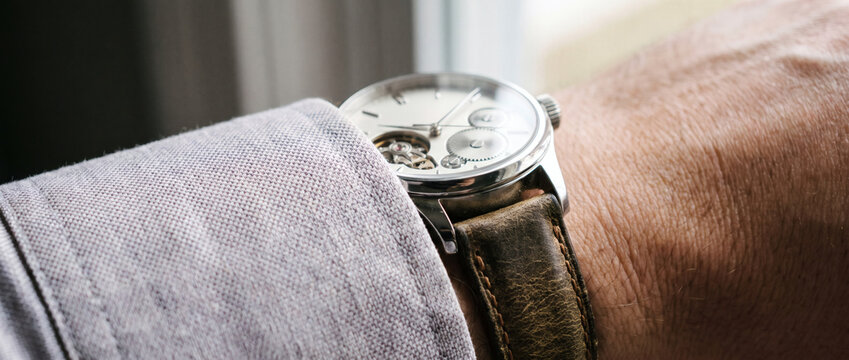 A classic mechanical wristwatch with exposed movement worn on a wrist, partially covered by a light shirt sleeve.