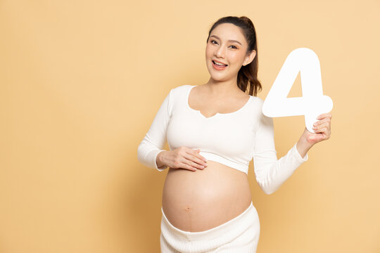 Pregnant Asian woman holding a number 4 sign. Happy mother four months or fourth trimester, showing bump. Maternity, healthcare concept isolated on brown background