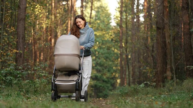 Young smiling mother pushing a baby stroller and walking on a path in a picturesque autumn forest. Lovely woman enjoying a stroll with her infant son through the sunlit woods