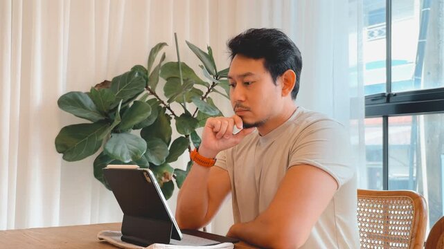Thoughtful Asian man concentrating on his digital tablet at a wooden table indoors.