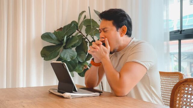 Tired Asian Man Yawning and Stretching While Working Remotely on a Tablet at Home