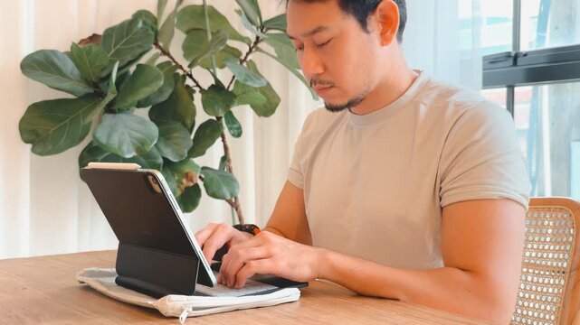 Focused Asian man working remotely on a tablet with a keyboard at a wooden desk.
