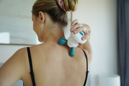 Young woman receiving neck and shoulder massage from handheld massager while seated on bed in bright bedroom