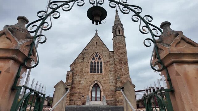 Slow motion view of the historic sandstone facade and gothic spire of St Patrick's Catholic Church in Stawell, Victoria, Australia framed by an wrought-iron gate archway. A heritage landmark.