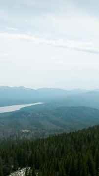 Wide aerial-style landscape view of Zyuratkul National Park with conifer forest, rocky slope, distant mountain ridges and a long lake under a cloudy sky, Russia